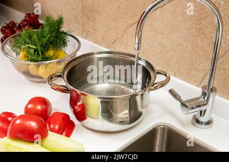 Das Wasser wird in einen Topf auf dem Tisch neben dem Spülbecken gezogen und gegartes Gemüse Stockfoto