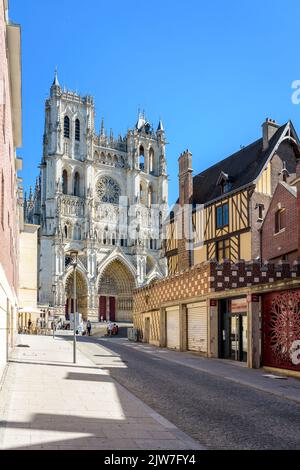 Gotische Fassade der Kathedrale Notre-Dame d'Amiens von einer schrägen gepflasterten Straße mit einem alten Fachwerkhaus an einem sonnigen Sommertag aus gesehen. Stockfoto