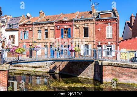 Typische Backsteinhäuser säumen den Fluss Somme im historischen Viertel Saint-Leu in Amiens, Frankreich, mit einer Fußgängerbrücke an einem sonnigen Sommertag. Stockfoto