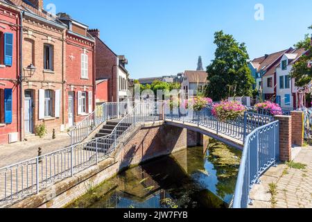 Typische Backsteinhäuser säumen den Fluss Somme im historischen Viertel Saint-Leu in Amiens, Frankreich, mit einer Fußgängerbrücke an einem sonnigen Sommertag. Stockfoto