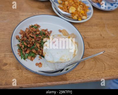 Pad Krapow MOO - Thailändisches Basilikum aus Schweinefleisch mit Reis und gebratenem Ei im Hintergrund. Thailändische Küche Gericht mit gehacktem Schweinefleisch, Basilikum, Soja und Austernsaucen. Thai Food Co Stockfoto