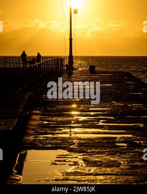 Der leuchtend gelbe Sonnenuntergang scheint auf dem Porthleven Pier, erstaunliche Reflexionen in den Pfützen vom Sonnenuntergang. Stockfoto