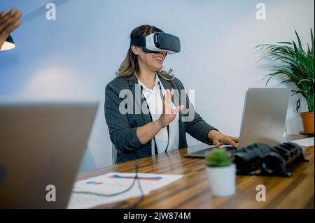 Glückliche weibliche Angestellte in VR-Brille, die im Büro auf dem Laptop tippt Stockfoto