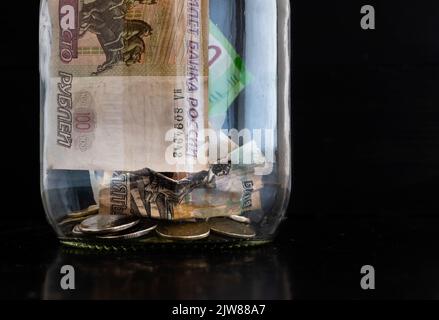 Zerknitterte Papierbanknoten russischer Rubel und Metallmünzen in einem Glasglas in Nahaufnahme auf schwarzem verschwommenem Hintergrund Stockfoto