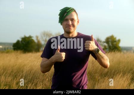 Junger glücklicher Mann mit grünen Dreadlocks, der ähnliche Gesten zeigt und am Vorabend des Sonnenuntergangs in der Natur steht. Positiver Hipster zeigt Daumen nach oben und schaut auf die Kamera Stockfoto