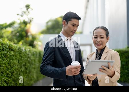 Erfolgreicher asiatischer Geschäftsmann und gealterte Geschäftsfrau, die vor dem Gebäude mit einem Tablet diskutieren Stockfoto