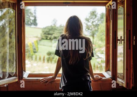 Rückansicht einer langen, dunkelhaarigen Frau in T-Shirt mit Blick aus dem Fenster und Blick auf die Landschaft mit rustikalem Naturbaumblick Stockfoto