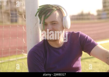 Junger Mann mit kabellosen Kopfhörern, der Musik hört, die Augen vor der strahlenden Sonne schließt, auf dem Fußballfeld sitzt und sich an die Tore lehnt Stockfoto