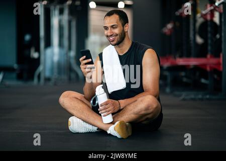 Happy Young Sportlicher Schwarzer Mann, Der Sich Im Fitnessstudio Mit Smartphone Und Wasser Ausruht Stockfoto