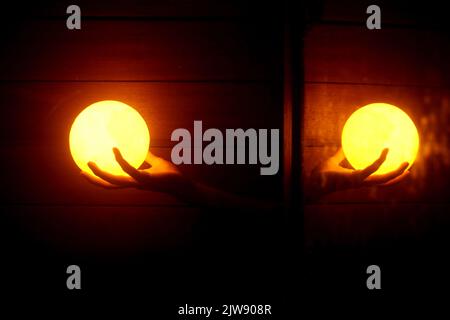 Person, die Balllampen in den Händen hält, mit hellem Spiegelschatten in der Dunkelheit. Doppelte gelbe Kugellampe in der Hand auf dunklem Hintergrund. Stockfoto