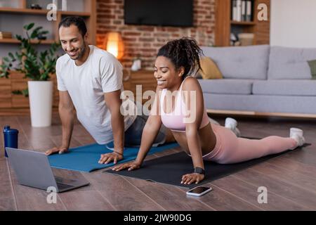 Ein frohes, junges schwarzes Paar in Sportswear macht Stretching oder praktiziert Yoga, schaut euch eine Videostunde auf dem Laptop auf der Matte an Stockfoto