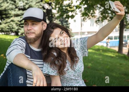 Ein Mann und ein Mädchen machen ein Selfie, das auf dem Gras im Park sitzt. Stockfoto