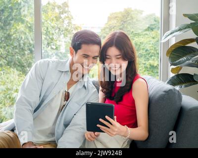 Glückliche junge schöne asiatische Frau und gutaussehende Mann lächeln zusammen, während sie auf Tablet-Bildschirm auf grauem Sofa in der Nähe des Glasfensters bei weißem Wohnzimmer ro Stockfoto