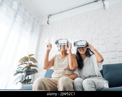 Asiatische junge Frau, glückliche Tochter und ihre Mutter, alte ältere Frau trägt VR-Brille, genießen 3D Spiel online zusammen im weißen Zimmer. Mutter und Tochter Stockfoto