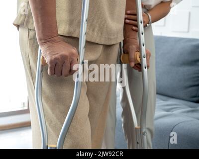 Nahaufnahme die Hände einer älteren Frau halten sich an Krücken, die zu Hause im Wohnzimmer stehen und versuchen zu gehen. Hilfe und Pflege des älteren Patienten beim Gehen mit Krücke Stockfoto
