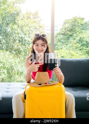 Junge, ziemlich glückliche asiatische Frau mit Sonnenbrille, die auf den Pass zeigt und mit einem Lächeln auf das Flugticket zeigt, sitzen auf dem Sofa in der Nähe des Strandhuts, gelber Koffer senkrecht Stockfoto