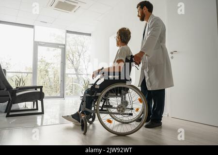 Professioneller männlicher Arzt, der weibliche Patientin auf dem Rollstuhl in der Medizinklinikhalle trägt Stockfoto