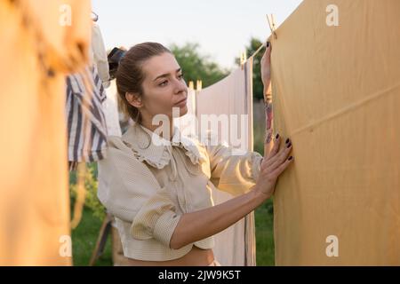 Wäschereitag. Eine Frau hängt Bettwäsche und Handtücher auf einem Baum im Hof eines Dorfhauses. Sommerhaus-Konzept. Stockfoto