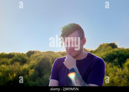 Ein junger, nachdenklicher Mann mit grünen Dreadlocks in einer Sonnenbrille schaut weg, kratzt sich mit dem Finger am Kinn und steht am Sonnenuntergang auf dem Hügel Stockfoto