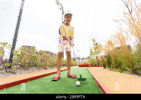 Nettes Mädchen im Vorschulalter, das mit der Familie Minigolf spielt. Glückliches Kind, das Spaß mit Aktivitäten im Freien hat. Sommersport für Kinder und Erwachsene, im Freien Stockfoto