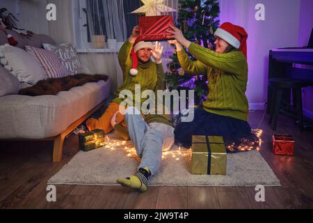 Glückliche Familie gibt Geschenke am Weihnachtsbaum im nächtlichen Licht der Girlande. Komfort am Silvesterabend im Wohnzimmer am Abend Stockfoto