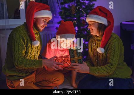 Glückliche Familie gibt Geschenke am Weihnachtsbaum im nächtlichen Licht der Girlande. Komfort am Silvesterabend im Wohnzimmer am Abend Stockfoto