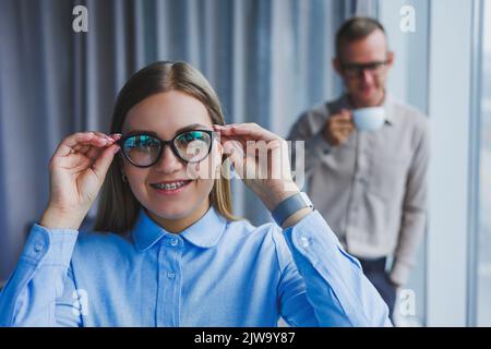 Mehrere freundliche Kollegen stehen am Bürofenster. Eine Geschäftsfrau in einer Brille steht auf dem Hintergrund eines Mannes in einem Hemd. Selektiver FOC Stockfoto