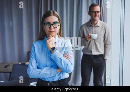 Mehrere freundliche Kollegen stehen am Bürofenster. Eine Geschäftsfrau in einer Brille steht auf dem Hintergrund eines Mannes in einem Hemd. Selektiver FOC Stockfoto