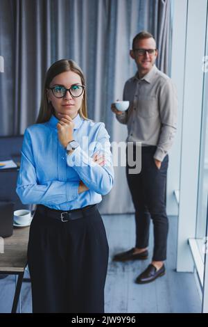 Mehrere freundliche Kollegen stehen am Bürofenster. Eine Geschäftsfrau in einer Brille steht auf dem Hintergrund eines Mannes in einem Hemd. Selektiver FOC Stockfoto