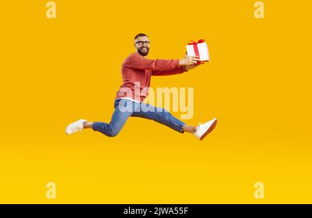 Glücklicher Mann hält Geschenkbox und springt in Luft isoliert auf orange gelben Hintergrund Stockfoto