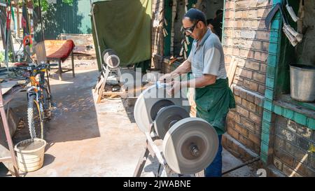 Osch, Kirgisistan - 2022. Mai: Mann, der in einer örtlichen Schmiede mit Funkenfeuerwerk ein Messer mit einer Mühle schärft. Arbeiter, die Schleifgeräte verwenden. Stockfoto