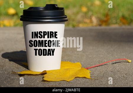 Auf einem gelben Ahornblatt steht eine Tasse Kaffee, auf der geschrieben steht - inspirieren Sie heute jemanden Stockfoto