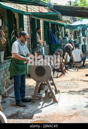 Osch, Kirgisistan - 2022. Mai: Mann, der in einer örtlichen Schmiede mit Funkenfeuerwerk ein Messer mit einer Mühle schärft. Arbeiter, die Schleifgeräte verwenden. Stockfoto