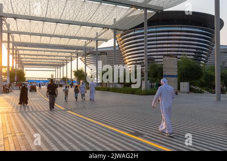 VAE, Dubai - 30. November 2021: Ausstellung EXPO 2020. Menschen gehen zwischen Pavillons entlang. Ausstellungshalle Mobilität in den Abendstrahlen des Sonnenuntergangs. Stockfoto