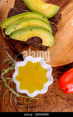 Olivenöl in weißer Tasse, rote Tomaten, Roggenbrot mit Avocado auf Holzschneidebrett. Stockfoto