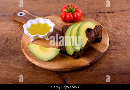 Olivenöl in weißer Tasse, rote Tomaten, Roggenbrot mit Avocado auf Holzschneidebrett. Stockfoto