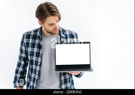 Ein moderner, gutaussehender, selbstbewusster Kaukasier, der auf einem weißen, isolierten Hintergrund steht, hält einen offenen Laptop, zeigt einen leeren weißen Bildschirm für Werbung oder Präsentation und sieht ihn an Stockfoto