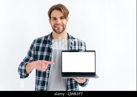 Stilvoller, gutaussehender, selbstbewusster Kaukasier, der auf einem weißen, isolierten Hintergrund steht, hält einen offenen Laptop, zeigt einen leeren weißen Bildschirm für Werbung oder Präsentation, zeigt den Finger darauf, lächelt Stockfoto