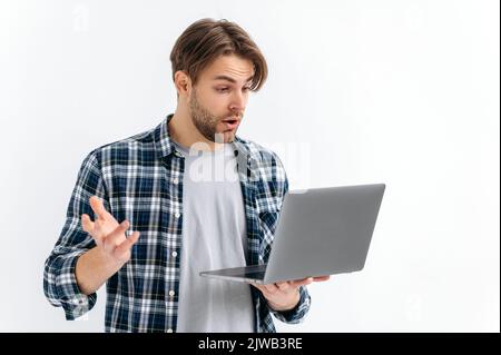 Schockiert verwirrt verwirrt verwirrt kaukasischen stilvollen jungen Mann, auf einem weißen isolierten Hintergrund stehend, hält einen offenen Laptop, überrascht schaut auf den Bildschirm, unerwartete Nachrichten sehen, Nachricht, gestikulierend mit der Hand Stockfoto