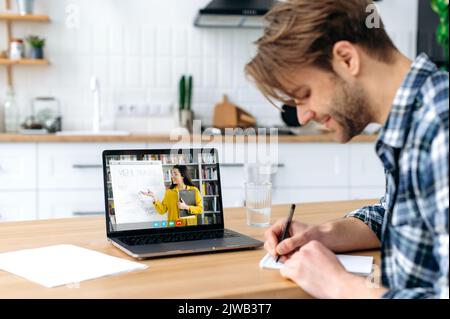 Online-Lernen. Seitenansicht eines smarten positiven kaukasischen Studenten, der zu Hause in einer Küche mit Laptop sitzt, einem Online-Vortrag zuhört, auf einem Bildschirm zeigt die kaukasische Lehrerin einer Schülerin aus der Ferne Stockfoto