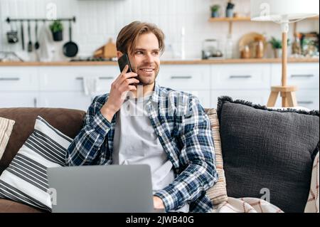 Glücklicher attraktiver moderner Kaukasischer, der während der Verwendung eines Laptops auf dem Handy spricht, zu Hause auf einem Sofa sitzt, ein angenehmes Gespräch mit einem Freund oder Kollegen führt, schaut weg und lächelt Stockfoto