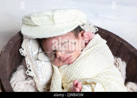 Neugeborener netter Junge mit Handtuch und Mütze bedeckt schlafen in hölzernen runden Schüssel dekorative kleine Requisiten und Blumen Stockfoto