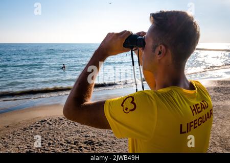 Rettungsschwimmer am Strand mit Blick durch ein Fernglas. Rückansicht des hübschen brünette männlichen Rettungsschwimmers mit gelbem Hemd am Strand im Sommer. Stockfoto