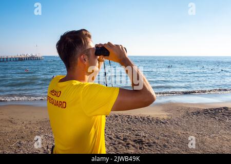 Rettungsschwimmer am Strand mit Blick durch ein Fernglas. Rückansicht des hübschen brünette männlichen Rettungsschwimmers mit gelbem Hemd am Strand im Sommer. Stockfoto