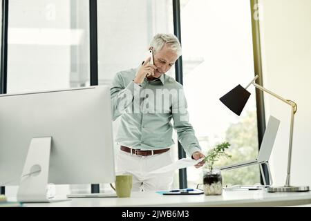 Ja, der Vertrag sieht perfekt aus. Ein reifer Geschäftsmann, der am Telefon telefoniert, während er an seinem Schreibtisch in einem Büro steht. Stockfoto