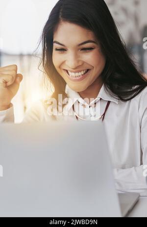 Der Erfolg kommt immer weiter: Eine junge Geschäftsfrau, die im Büro an einem Laptop arbeitet und eine Faustpumpe macht. Stockfoto