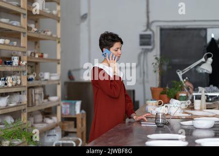 Junge Frau Keramik Studio-Manager mit Telefongespräch und Laptop, Töpferei Shop Besitzer bei der Arbeit Stockfoto