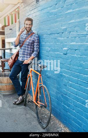 Ich hüpfe jetzt auf mein Fahrrad. Ein hübscher junger Mann, der sich mit seinem Fahrrad auf einem Handy an eine Wand lehnt. Stockfoto