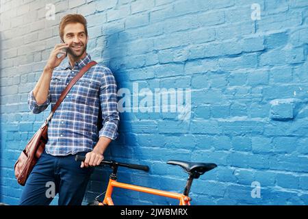 Ich werde bald Ihren Weg Rollen. Ein hübscher junger Mann, der sich mit seinem Fahrrad auf einem Handy an einer Wand lehnt. Stockfoto