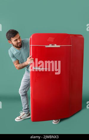 Cooler tätowierte junge Mann, der aus dem roten Kühlschrank auf grünem Hintergrund kommt Stockfoto